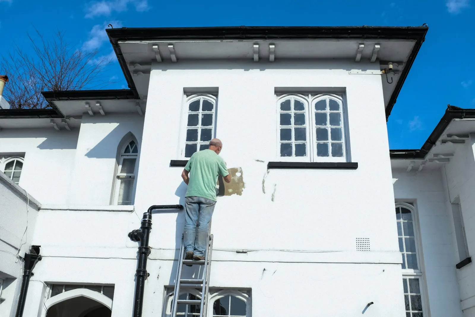 Un homme debout dans une échelle repeint le mur extérieur blanc d’une maison à étage, sous un ciel bleu