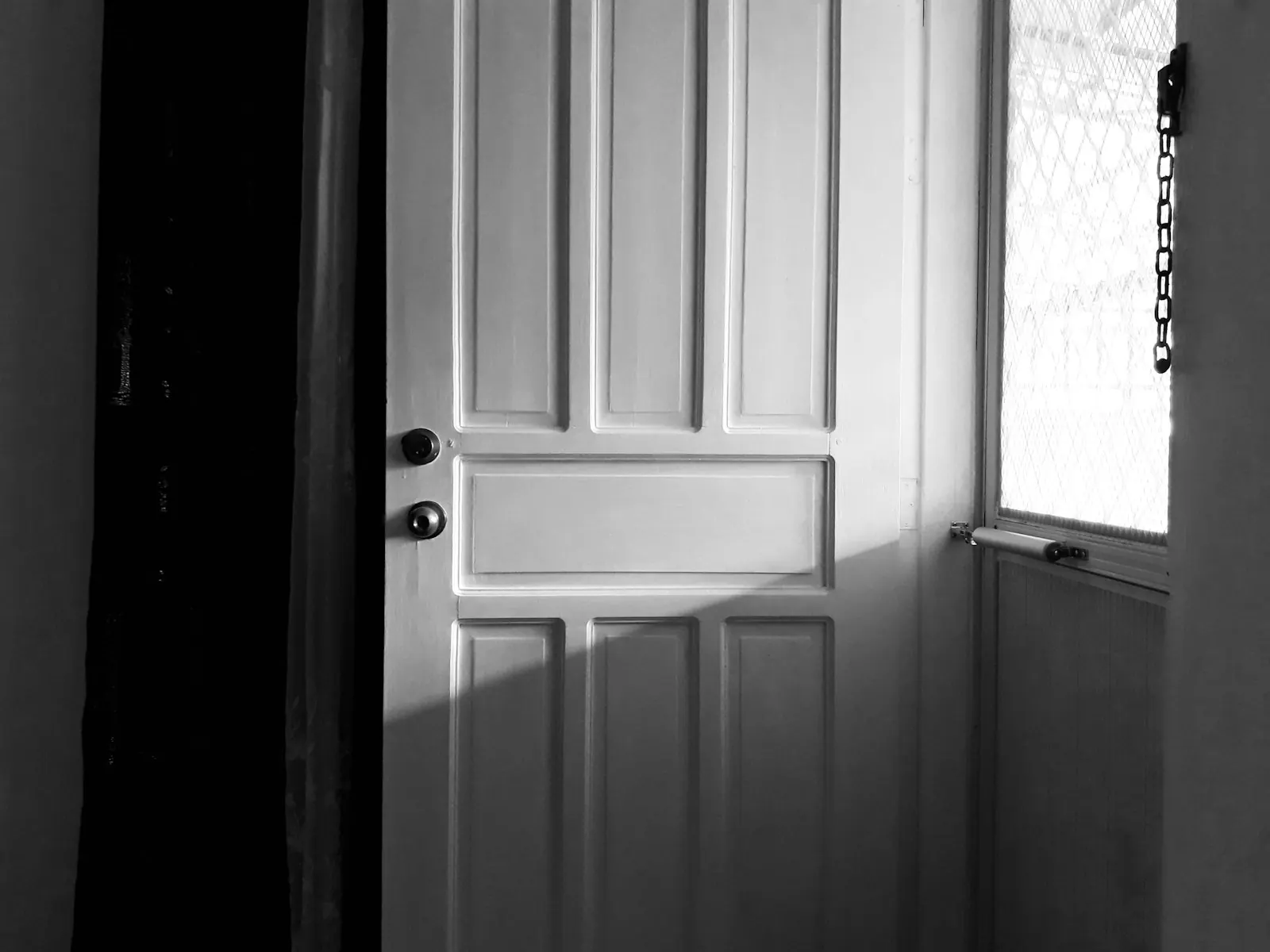 Une porte intérieure blanche à panneaux, entrouverte, avec une poignée noire, photographiée en noir et blanc