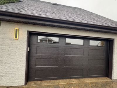 Porte de garage double brune imitation bois avec trois fenêtres rectangulaires, sur une maison au crépi blanc